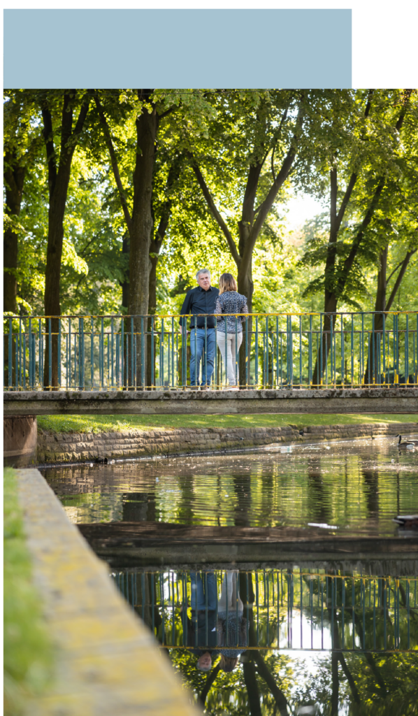 Das Bild zeigt eine Frau und einen Mann, die sich unterhalten, in einem Park. Gemeinsam stehen sie auf einer Brücke.Im Hintergrund sind viele grüne Bäume zu sehen. Im Vordergrund spiegelt sich das Bild in einem Fluss.