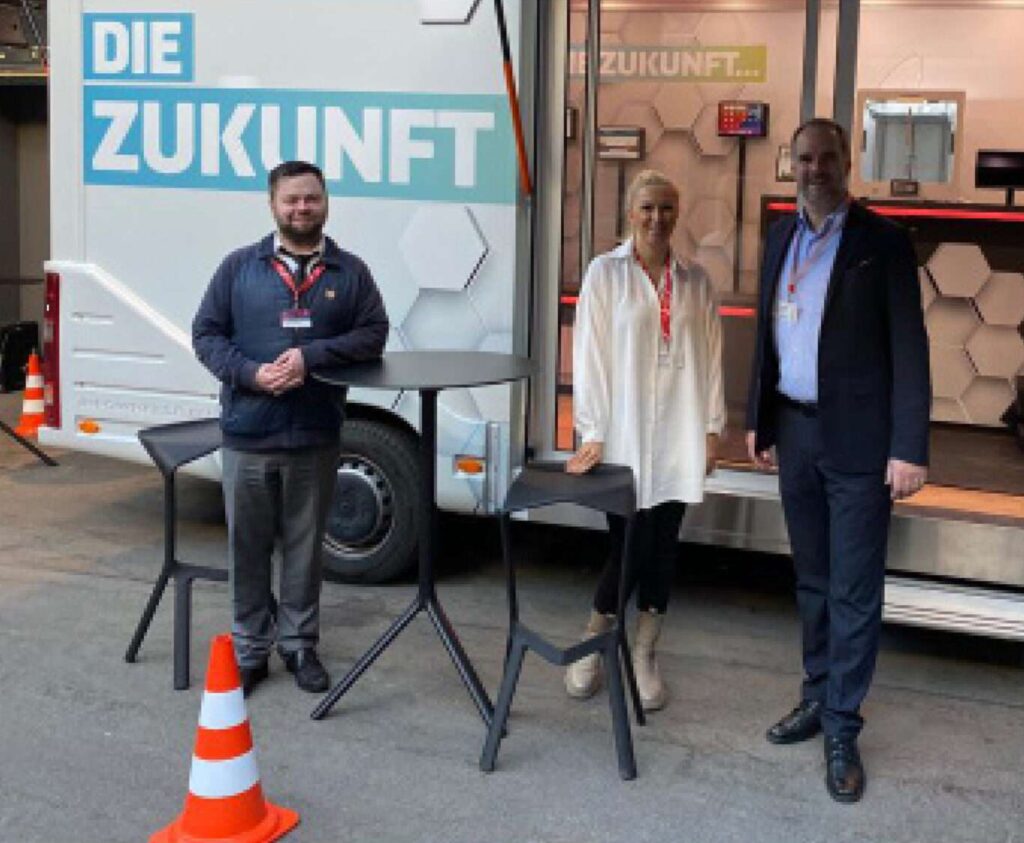 Bild von drei Personen, die vor einem Verkaufsstand (Wohnmobil) stehen.