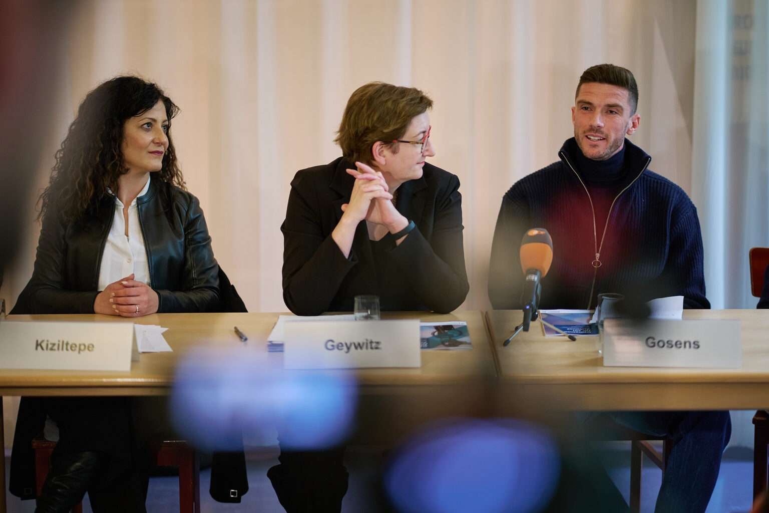 Der Fußballspieler Robin Gosens sitzt gemeinsam mit zwei weiteren Frauen an einem Tisch bei der Pressekonferenz. Er ist gerade am sprechen. Die anderen beiden Frauen links von ihm schauen ihn an und hören gespannt zu.