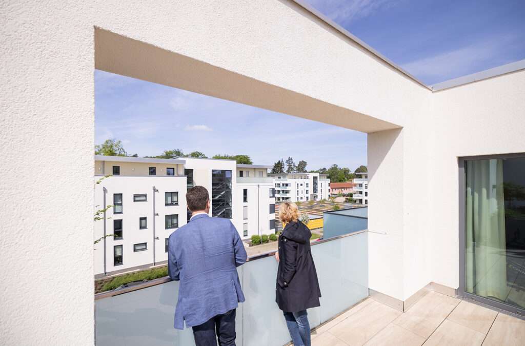 Das Bild zeigt eine Frau und einen Mann, die gemeinsam auf einer Dachterasse stehen und in die Ferne auf Gebäudekomplexe schauen.